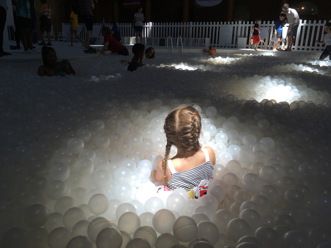 a large ball pit full of translucent white balls, with a kid in braids and a dress in the foreground of the ball pit, other people in the distance of the ball pit, and white beach umbrellas and lounge chairs at the far edge